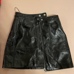 Svart kjol från Mango - En svart kjol från Mango i storlek S. Kjolen har en glansig yta och är utrustad med en dragkedja framtill samt två knappar i midjan. Den har också två fickor framtill. Skriv gärna om ni har frågor 💕