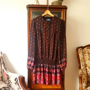  - Otrolig klänning/tunika i tunt retro-mönstrat tyg, har även detaljer som band att knyta i vristen och har flera knappar i brons.  Kan avändas som klänning eller tunika men bör ha något under då tyget i vissa ljus uppfattas genomskinligt. (Om du vill)  Storlek 38. I fint skick. 