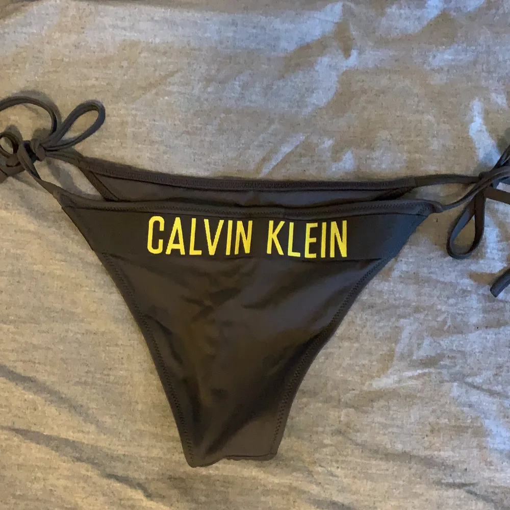 En mörkgrå Calvin Klein bikini underdel med gul text på baksidan. Knutarna kan man lösa up och justera. Använd några få gånger.. Övrigt.