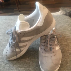  - Säljer mina sneakers som jag köpte förra våren men knappt använt. Ett par ljusgråa Adidas Gazelle med vita ränder, gazelle skrivet i guld, storlek 40 US 8, UK 6,5. Jättefint skick! Endast använda en gång...