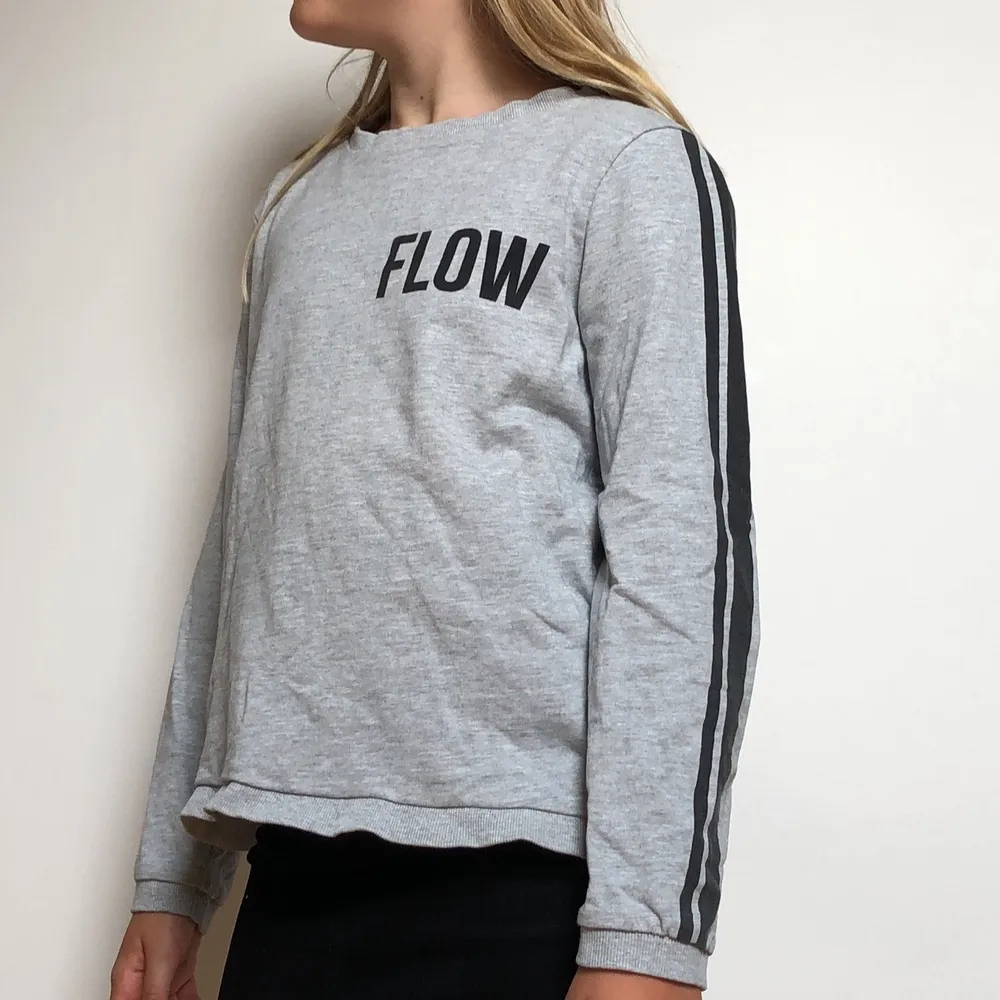 Ljus grå långärmad tröja med svarta sträck på armarna. Sedan står det ”Flow” på framsidan och på baksidan finns ett ”v” format hål.. Tröjor & Koftor.