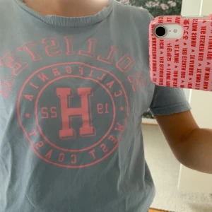 Hollister t-shirt  - Blå Hollister t-shirt med rosa tryck och slitningar vid halsen. Köparen står för frakten 