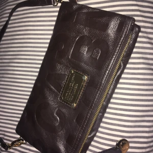  - En väska från Marc jacobs  som är gjort av skin som är brunt och inne i den är det svart vit text med Marc Jacobs man kan fälla ut den så den blir större