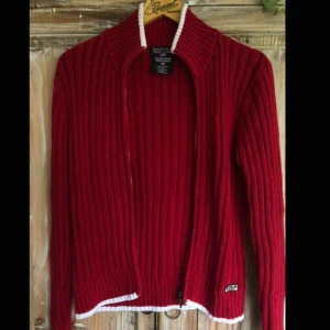  - Stickad röd Polo Ralph Lauren kofta med dragkedja i fram. Vita accenter längst ner, samt vid kragen och vid ärmarna. Väldigt fint skick, nästan oanvänd.