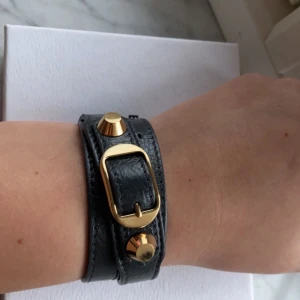 Balenciaga armband  - Svart Balenciaga armband med guldiga detaljer i nyskick, såklart äkta!! Använt max 3 gånger och har inga slitage. Köpt på Nathalie Schuterman som en present så kvitto finns tyvärr inte kvar. Box och dustbag följer med. Kontakta mig för flera bilder. Kan mötas upp och frakta eller byta mot valentino armband i nyskick.