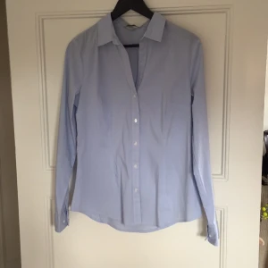  - Ljusblå skjorta med smala vita ränder från H&M. Skjortan är insvängd vid midjan och är sparsamt använd. 