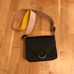 Väska från hm - Liten svart väska från hm. Bandet är gult på ena sidan och rosa på andra. Väskan är som ny och bara använd någon enstaka gång.