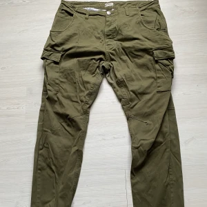 Militärgröna cargo pants - Militärgröna cargo pants från Lager 157 med fickor fram, bak och på benen. Elastisk mudd längst ner på benet. Ingen dragkedja i gylfen utan man knäpper endast med gömda knappar:) Frakt: 80kr