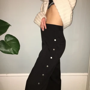  - Oanvända, trendiga svarta byxor med knappar längs sidorna.