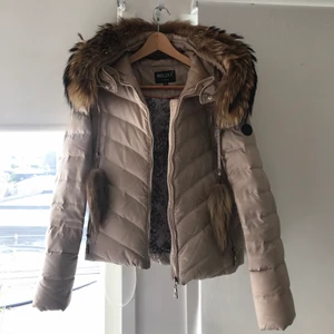 Hollies jacka - En beige Hollies jacka i storlek 34. Skulle säga att den är i använt skick och har en defekt på ena armen men det syns inte när man har jackan på sig. Pälsen är äkta men den är avtagbar, samma med ”tussarna” som hänger