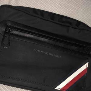 En Tommy Hilfiger väska i färgen svart, jag säljer den helt ny och oanvänd. Det är en ganska stor väska med tre olika fickor, den är cirka 20 cm lång och 14 cm hög. 