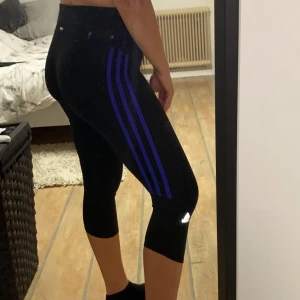 Adidas träningstights, korta - Svarta adidas träningstights i super skönt material med lila/blåa streck vid sidorna. kortare variant som går ner strax över knäna. Även en ficka vid sidan av höften (bild 2). Frakt tillkommer💙🖤