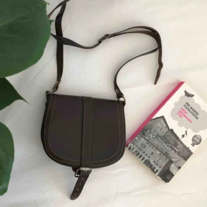  - Gammal vintage väska i mörkbrunt läder, i fint skick. Se storleken i förhållande till en vanlig pocketbok på första bilden. Axelbandslängd (cm) 98-118. Säljes pga att den inte används längre. Kan skickas, då står köparen för fraktkostnaderna. Har swish.