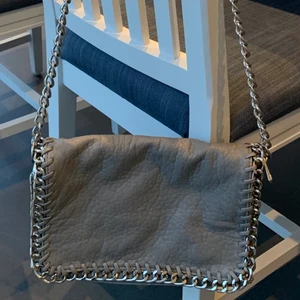 Tiamo, modell Priscilla, grå axelremsväska med kantad silverlänk - Tiamo, modell Priscilla, grå axelremsväska med kantad silverlänk i jättefint skick. Ett fack med dragkedja på baksidan och ett fack med dragkedja på insidan. Avtagbar axellänk gör att den kan bäras även som en clutch. Axellänk 105 cm. Längd 28 cm, höjd 18 cm, bredd 5 cm. Inköpt på Scorett. 150kr/eller bud.
