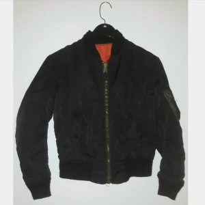  - Svart bomberjacka med orange foder. 2 yttre fickor och 2 innerfickor, alla stängs med tryckknapp. tryckknappen till innerfickan på ena sidan är trasig (se 3e bild) annars bra skick.  Lite noppriga muddar kanske, kan ta bättre bild.