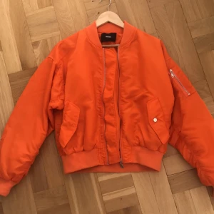  - Bomberjacka i stark orange färg (lyser lite mer orange i verkligheten). Superbra skick förutom att en knapp lossnat (se sista bilden). Finns en extra knapp i fickan som går att sy fast!