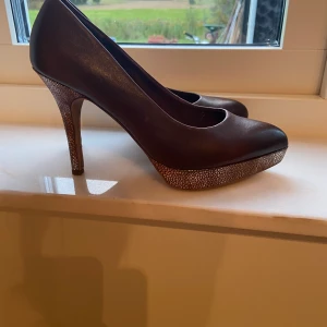 Pumps från Tamaris - Snygga pumps från Tamaris strl 37. Fint skick och endast använda 1-2 ggr. Brunt läder med guldiga klackar. Klackhöjd 10cm inkl 2 cm plattå i tån. Orginal kartong finns kvar. Nypris 795kr. Katt finns i hemmet. Skickas med postens skicka lätt, frakt ingår i priset betalas med swish. 