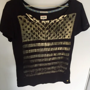 Guld/svart Tommy Hilfiger-T-shirt - Jättesnygg Tommy Hilfiger-T-shirt som hängt i garderoben nått år. Använd ca 5 gånger. Kommer den mer till användning hos dig? ;)
