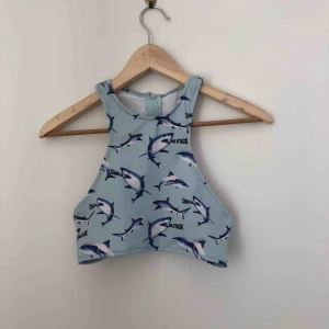  - Blå bikiniöverdel med brottar-rygg och dragkedja. Motiv med hajar och texten ”shark attack”. Sparsamt använd och i bra skick!  Kan mötas längst roslagsbanan eller i stockholm, annars står du för frakt.