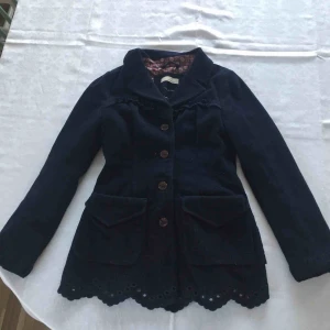  - Mörkblå vinter/höst/vår jacka från Odd Molly. I fint skick. Hel utom en söm i innerfordret (se bild 3). Jackan i ull och cashmere. Detaljer med rosett bak och brodyr och spets längs nedre kanten. Broderade blommor i fickorna och fjärilar på knappar.