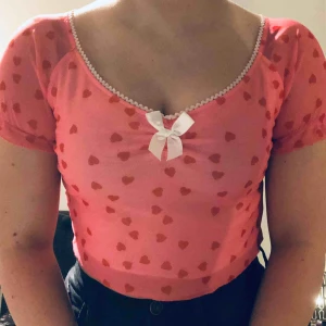  - Supersöt rosa topp med röda hjärtan och vit rosett framtill! Tröjan är i mesh men har ett lager vitt tyg under. Liten ”ruffle” på ärmarna och nedtill. Frakt ingår❤️💗