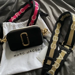 Marc Jacobs väska snapshot  - Säljer denna fina väskan, med orginalbandet (de rosa) + ett jag köpt till senare (guld). Köpte väskan för 3299kr, samt guld bandet för 999kr. Säljer nu väskan + de två banden för 900kr. Man får även med dustbag och äkthetsbevis. 