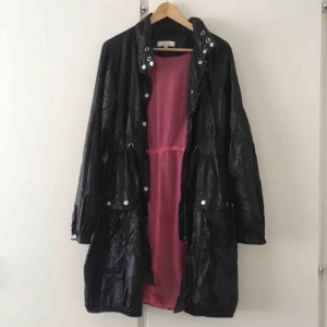  - Lång svart jacka med ros nätfoder som ger bra ventilation. Har luva som rullas diskret in in kragen. 