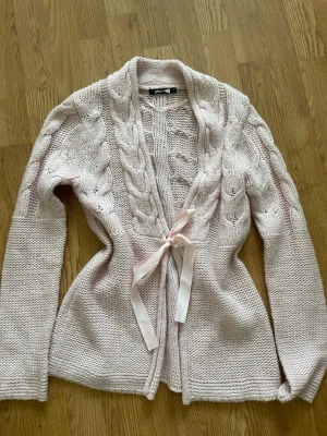 Ljusrosa stickad kofta med rosett - Supersöt ljusrosa stickad kofta från gina tricot med flätstickade detaljer och öppen front. Koftan har en fin satinrosett i midjan och långa ärmar.🫶