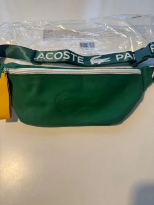 Grön magväska från Lacoste - Snygg grön magväska från Lacoste med vitt blixtlås och justerbart band där det står 'Lacoste Paris'. Väskan har en gul dragkedjehållare och är tillverkad i ett slätt material med Lacoste-loggan präglad på framsidan. Perfekt för att bära det viktigaste.