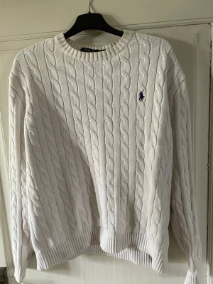 Vit kabelstickad tröja Polo Ralph Lauren - Kabelstickad vit tröja från Polo Ralph Lauren med rund halsringning och ribbade muddar. Klassisk logga i marinblått broderad på bröstet. Tröjan har en avslappnad passform och är perfekt för lager-på-lager. Materialet känns mjukt och bekvämt.