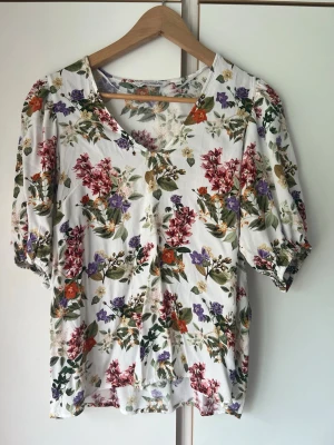 Blommig vit blus med puffärm - Söt blus med vit botten och färgglatt blommönster i rött, lila, orange och grönt. Blusen har v-ringning och puffiga korta ärmar med resår vid ärmslut. Perfekt för en somrig och fräsch look.