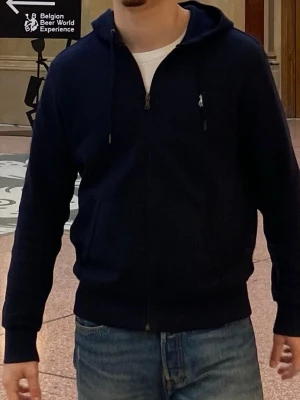 Mörkblå hoodie från Polo Ralph Lauren - Snygg mörkblå hoodie från Polo Ralph Lauren med dragkedja framtill och vit broderad logga på bröstet. Hoodien har huva med dragsko och ribbade muddar vid ärmslut och nederkant. Perfekt till jeans för en clean och stilren look.