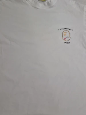 Vit t-shirt från A Bathing Ape - Vit t-shirt från A Bathing Ape med broderad logga och texten 'A Bathing Ape Japan' på bröstet. På ryggen finns ett stort tryck med apans ansikte, japanska städer och texten 'Ape Shall Never Kill Ape'. Klassisk passform och rund hals i mjuk bomull.