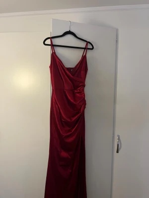 Röd långklänning med omlott - Elegant röd långklänning med omlottdesign och smala axelband. Klänningen har en v-ringad urringning och draperad detalj i midjan som ger en snygg siluett. Perfekt för speciella tillfällen där du vill sticka ut.