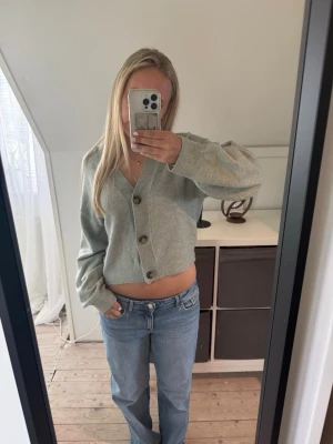Grå cropped kofta med knappar - Mysig grå kofta med v-ringning och stora bruna knappar framtill. Modellen är cropped och har breda ribbade muddar vid ärmslut och nederkant. Perfekt att slänga över en t-shirt eller linne för en chill look.