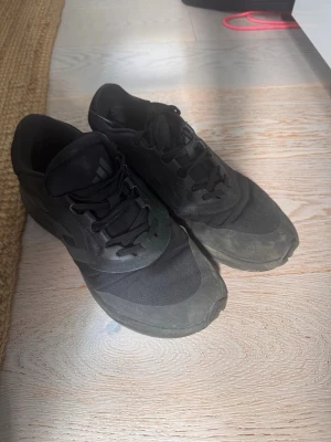 Svarta Adidas sneakers - Säljer ett par helsvarta Adidas sneakers med klassisk låg profil. Skorna har ovandel i textil och syntet, samt svarta snören och diskret Adidas-logga på plösen. Snygg och enkel design som passar till det mesta.