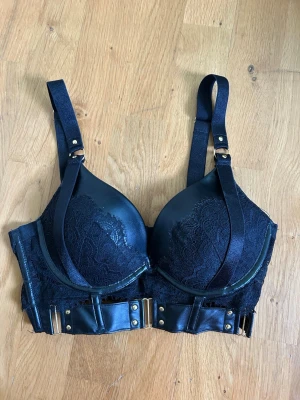 Svart Bygel-Bh - Hunkemöller - 🖤 Snygg svart bh med bygel, vadderade kupor och dekorativa band över bysten. Bh:n har spetsdetaljer, bredare band och guldfärgade metalldetaljer. Använd fåtalet gånger och i mycket fint skick. Inköpt på Hunkemöller. 