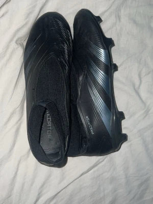 Adidas Predator svarta fotbollsskor - Säljer ett par svarta Adidas Predator fotbollsskor med strumpliknande skaft och mönstrad ovandel. Skorna har en modern, strömlinjeformad design utan snörning och fasta dobbar för bra grepp på planen. Tillverkade i syntetmaterial för lätt vikt och bra bollkontroll.