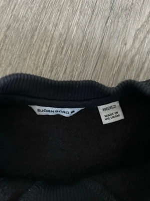 Svart sweatshirt från Björn Borg - Snygg svart sweatshirt från Björn Borg med logga i vitt på bröstet. Tröjan har rund halsringning, långa ärmar och ribbade muddar vid ärmslut och nederkant. Tillverkad i mjukt material som känns skönt mot huden.