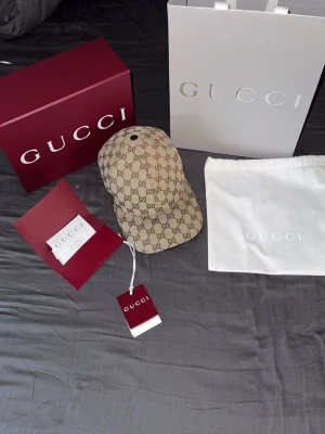 Beige GG Supreme keps från Gucci - Säljer en beige keps från Gucci med det klassiska GG Supreme-mönstret över hela kepsen. På sidan finns en grön och röd rand, typisk för Gucci. Kepsen har böjd skärm och justerbart band bak. Kommer med originalförpackning, dustbag och taggar.köpt i Danmark för 3500dk