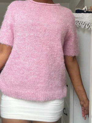 Ljusrosa stickad t-shirt - Mysig ljusrosa stickad t-shirt med korta ärmar och rund hals. Tröjan har en fluffig, mjuk struktur och avslutas med ribbstickade kanter. Perfekt för dig som gillar en söt och enkel look.