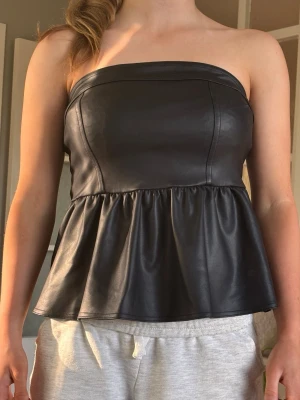 Svart axelbandslös peplum tubtopp - Snygg svart axelbandslös tubtopp i skinnimitation med peplumvolang. Toppen har en markerad söm framtill och dragkedja bak för enkel påklädning. Perfekt för att skapa en edgy look till jeans eller kjol.
