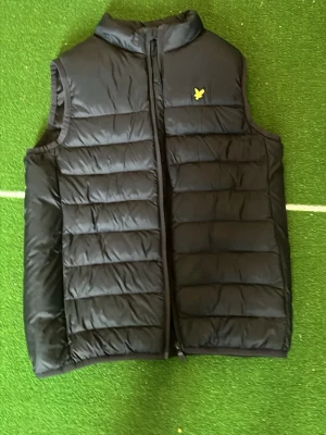 Svart dunväst från Lyle & Scott - Snygg svart dunväst från Lyle & Scott med gul logga på bröstet. Västen har inga skavanker och är bara använd under få tillfällen, fickor på sidan även. Perfekt att slänga över en hoodie eller tröja för extra värme.