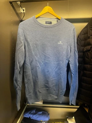 Blå rundhalsad tröja från Gant - Snygg blå rundhalsad tröja från Gant med diskret logga broderad på bröstet. Tröjan har en tunn stickning och ribbade muddar vid ärmslut och nederkant. Perfekt till jeans eller chinos för en clean och stilren look.