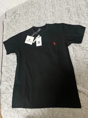Svart t-shirt  - Klassisk svart t-shirt med korta ärmar och rund hals. T-shirten har den ikoniska röda Polo-loggan broderad på bröstet och är tillverkad i mjuk bomull för en bekväm känsla. Perfekt basplagg till garderoben.