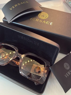 Versace rosa solglasögon med guldlogo - Snygga solglasögon från Versace med rektangulära, breda bågar i transparent rosa plast och  guldig Medusa-detalj på sidorna. Orginalptis 3100kr!!