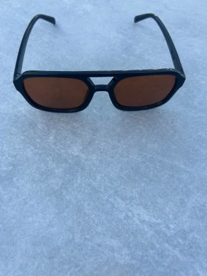 Svarta solglasögon med bruna glas - Snygga solglasögon med svart, kantig båge och bruna glas. Modellen har en modern, fyrkantig form och bred brygga. Perfekta för dig som vill ha en stilren accessoar med retrovibbar. Verkligen helt unika och perfekta till sommaren och soliga höstkvällar 