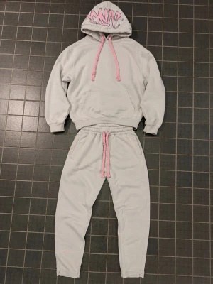 Äkta Gråa syna world tracksuit  - Mysiga grå syna world tracksuit från synaworldwideuk.com storlek m med breda rosa dragsnören och matchande hoodie. Hoodien har graffiti-inspirerat tryck på luvan och stor ficka framtill. Perfekt för chill dagar. Materialet är mjukt och skönt.