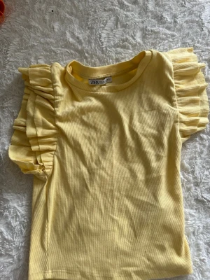 Gul ribbad topp med volangärm Zara - Supersöt ljusgul ribbad topp från Zara med volangärmar som ger en extra touch. Rund halsringning och mjukt material som känns skönt mot huden. Perfekt för dig som vill ha något färgglatt och trendigt i garderoben.