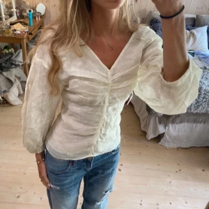 Vit v-ringad blus med puffärm - Säljer en vit blus med v-ringning och puffiga långärmar. Blusen har rynkade detaljer framtill och är tillverkad i ett tunt, lätt material som ger en luftig känsla. Perfekt att styla med jeans för en trendig look.